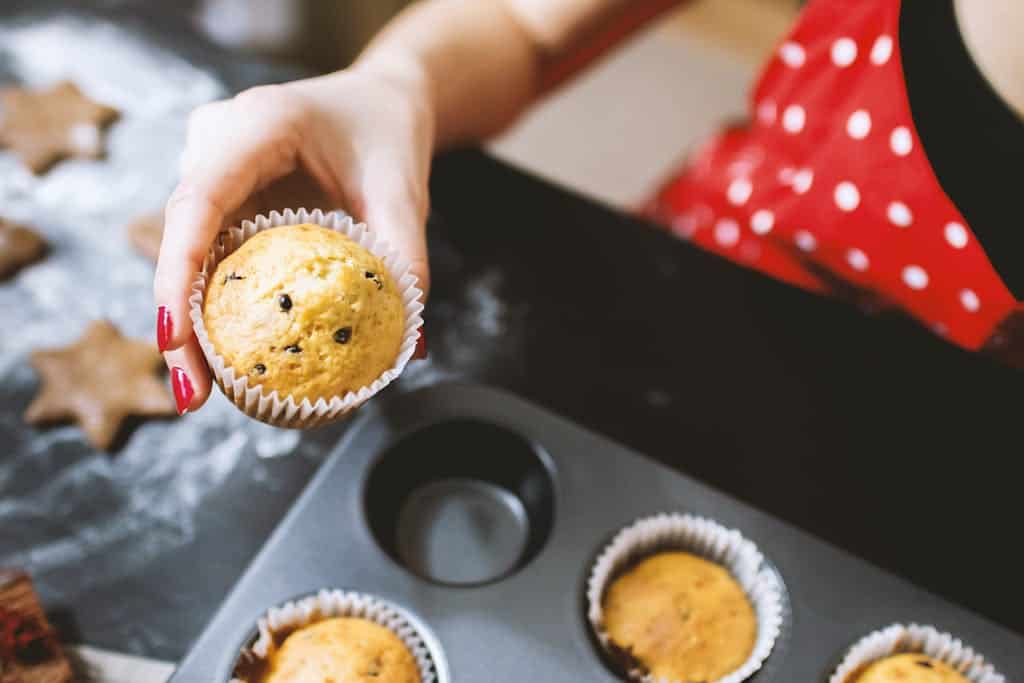 A person holding a chocolate chip cupcake fresh from the oven in a kitchen setting.