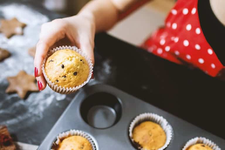 A person holding a chocolate chip cupcake fresh from the oven in a kitchen setting.